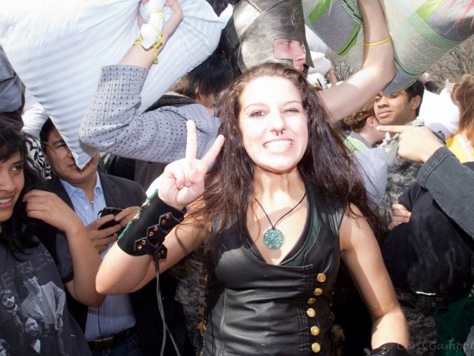 OLYMPUS DIGITAL CAMERA A woman smiles and flashes a peace sign amid a lively crowd at a pillow fight event.
