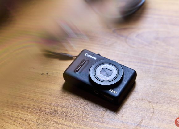 A black Canon S100 digital camera rests on a wooden surface with soft light and shadows.