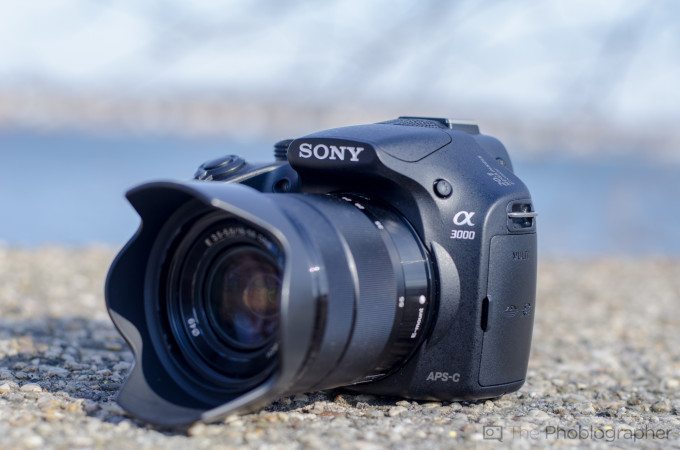 A black Sony Alpha a3000 camera with lens sits on a gravel surface, blurred background.