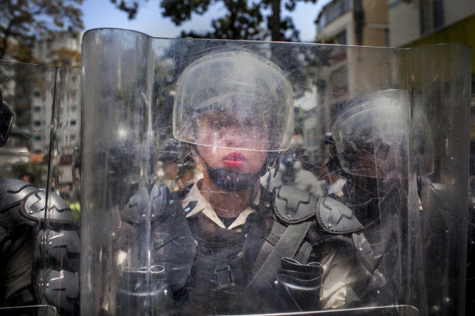 March 6th, 2014. Caracas, Venezuela. A National Police officer behind a riot shield is pushed backwards by a crush of demonstrators during the March of the Empty Pots, which coincided with International Women's Day.  A month into a wave of unrest that has spread across Venezuela, protests continued daily in the upper class enclave of Altamira, Caracas, as well as other parts of the city and country. Protesters are calling for solutions to a staggering rate of street crime, skyrocketing inflation, and a rash of food shortages that have led to long lines at grocery stores and lack of access to goods such as milk, sugar, and flour. While many large protests have been peaceful, the youth demonstrations each afternoon in Altamira are consistently characterized by exchanges of tear gas canisters and sometimes birdshot on the part of police, and rocks, molotov cocktails and other fireworks on the part of the protesters.