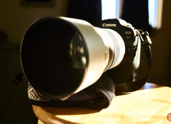 A Canon EOS R3 camera with a large white lens rests on a wooden surface. The iconic camera strap is visible, and the lens cap is off, revealing the pristine front glass element. The background is softly blurred, highlighting the sleek design of the Canon EOS R3.