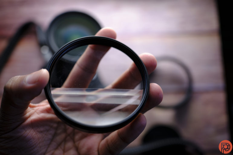 A hand holds a round camera lens filter with blurred camera gear in the background.