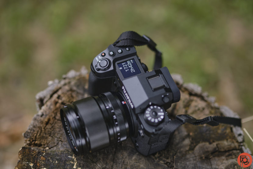 Phoblographer Readers Get a Special Perk When Buying a Fujifilm X-H2S Black mirrorless camera with a zoom lens resting on a tree stump, with blurred green background.