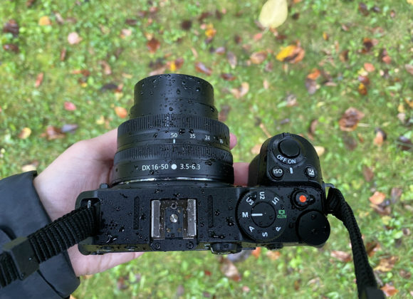 A hand holding a camera with raindrops on it, above grassy ground scattered with autumn leaves.