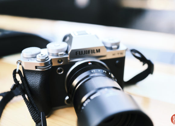 A Fujifilm digital camera with a zoom lens rests on a wooden table, with a blurred background.