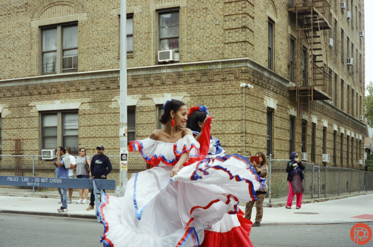 Two women in colorful traditional dresses dance on a city street, with onlookers and brick buildings behind them.