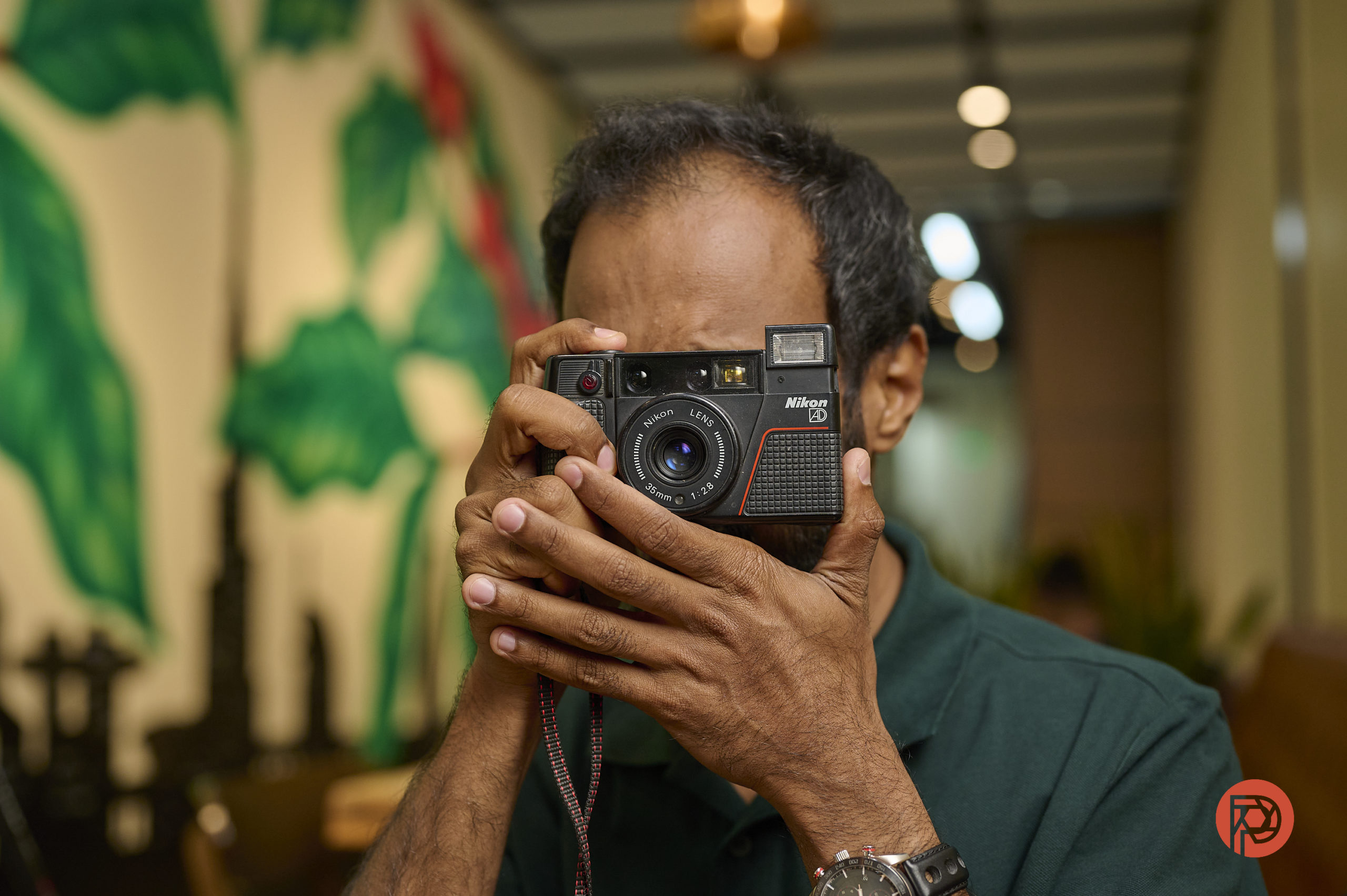 A person in a green shirt holding a Nikon camera up to their face, about to take a photo indoors.