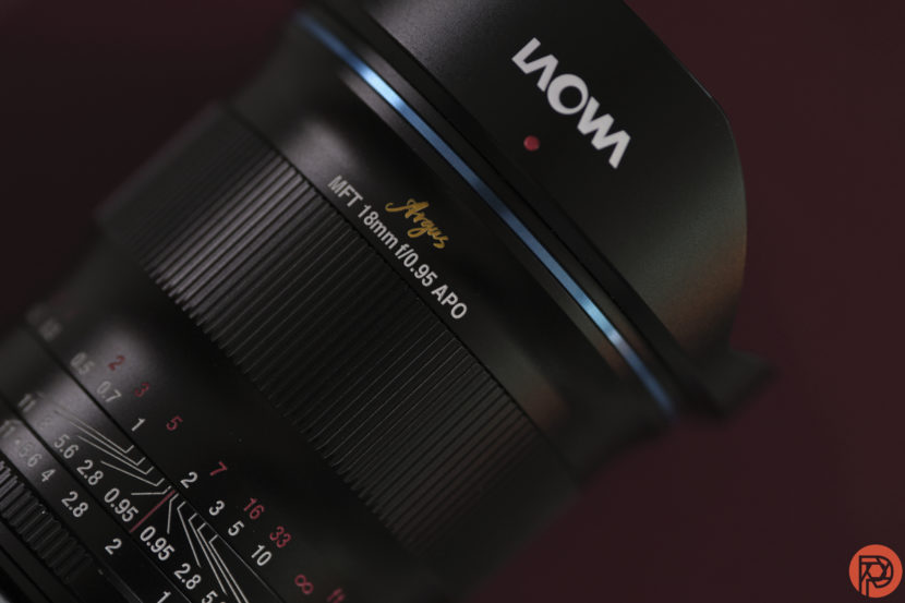 Close-up of a Laowa Argus MFT 18mm f/0.95 APO camera lens against a dark red background.
