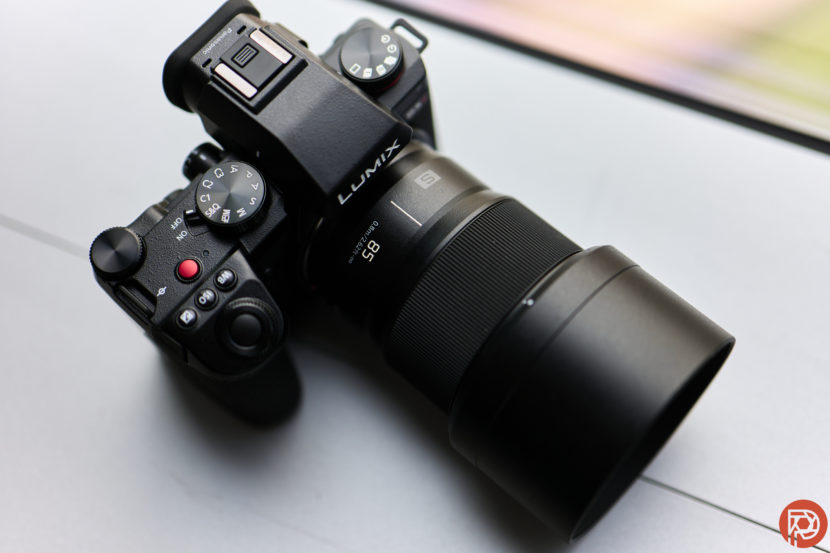 Black Lumix camera with an 85mm lens on a light gray surface.