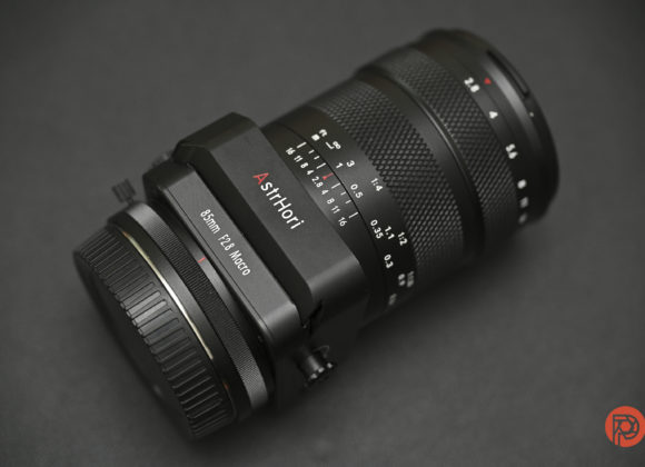 A close-up of a black AsriHori 85mm f/2.8 macro camera lens on a dark surface.