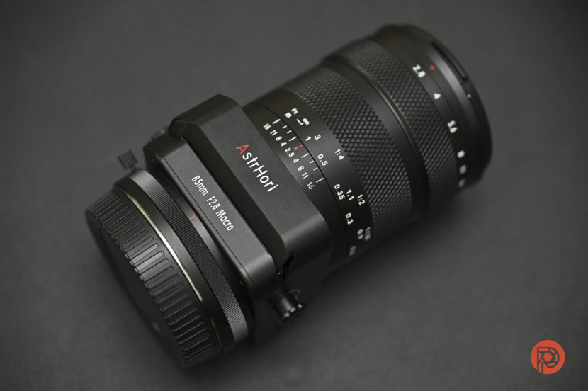 A close-up of a black AsriHori 85mm f/2.8 macro camera lens on a dark surface.