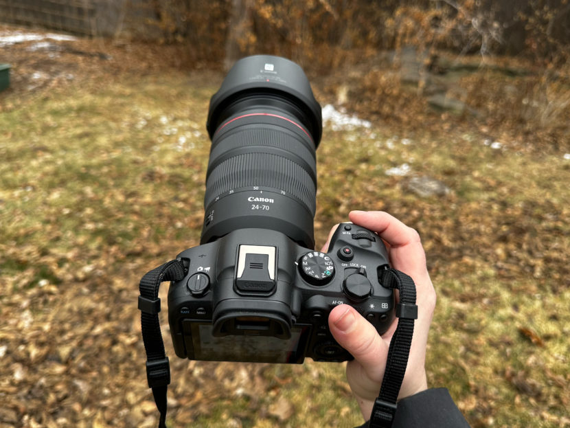 A hand holding a Canon camera with a 24-70mm lens outdoors, on brown grass with patches of snow.