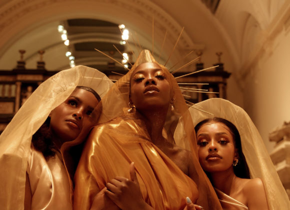 Three women in elegant gold and cream gowns pose under an archway. The central figure wears a radiant halo headpiece.