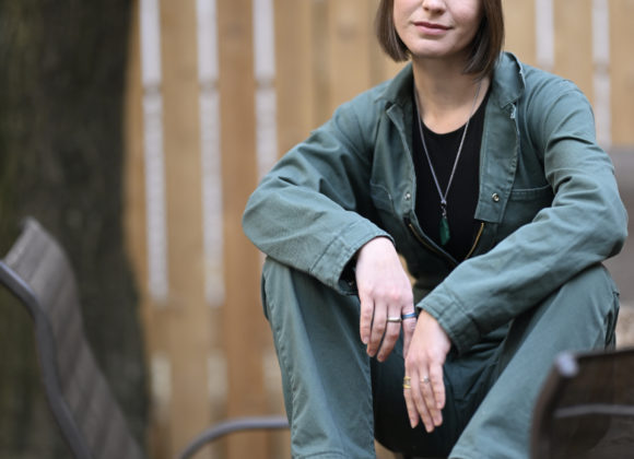A person in a green jumpsuit and orange beanie sits on a metal chair outdoors, smiling slightly.