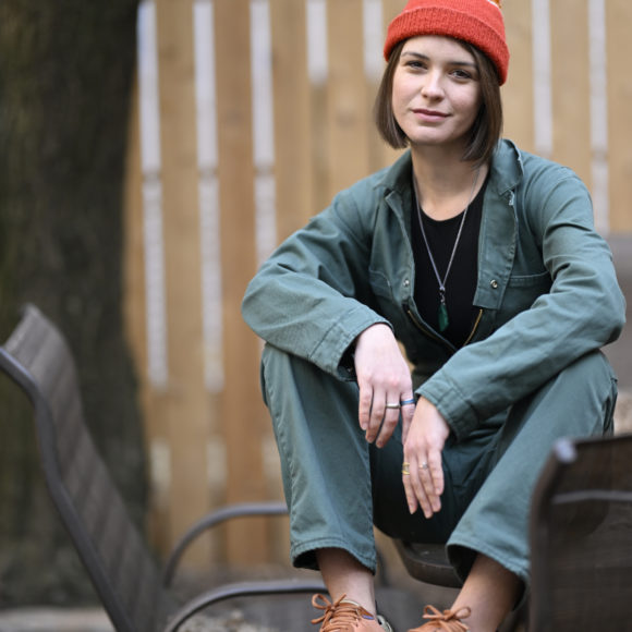 A person in a green jumpsuit and orange beanie sits on a metal chair outdoors, smiling slightly.