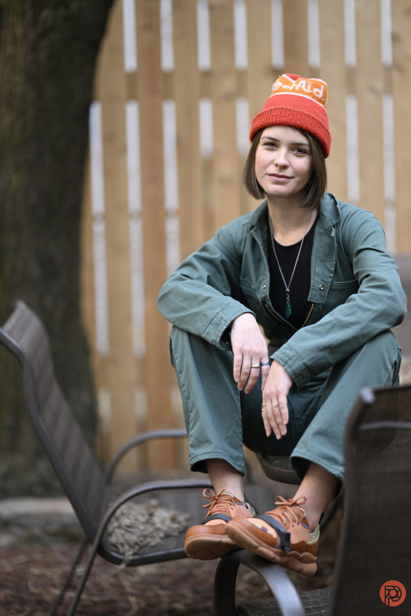 How to ACTUALLY be a Good Portrait Photographer A person in a green jumpsuit and orange beanie sits on a metal chair outdoors, smiling slightly.