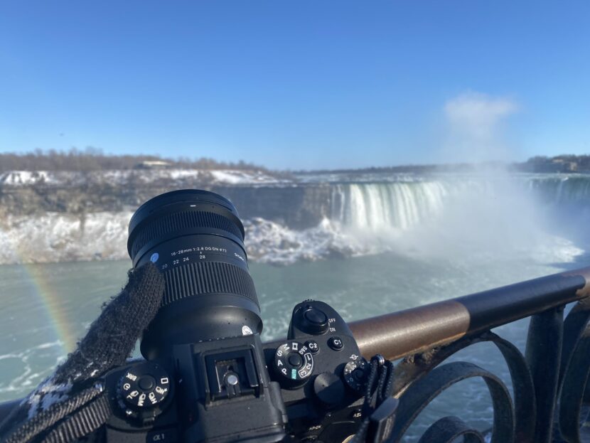 A camera on a railing captures Niagara Falls with a rainbow and blue sky in the background.