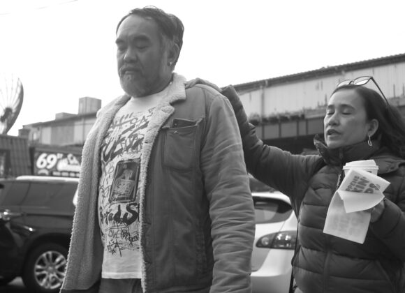 A man carrying shopping bags walks ahead as a woman with papers reaches toward his shoulder on a city street.