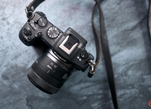 A Canon mirrorless camera with a zoom lens and strap resting on a textured blue surface.