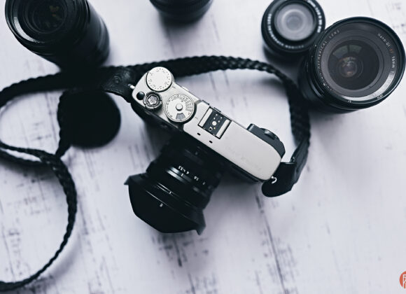 A silver camera with a strap surrounded by three camera lenses on a white wooden surface.