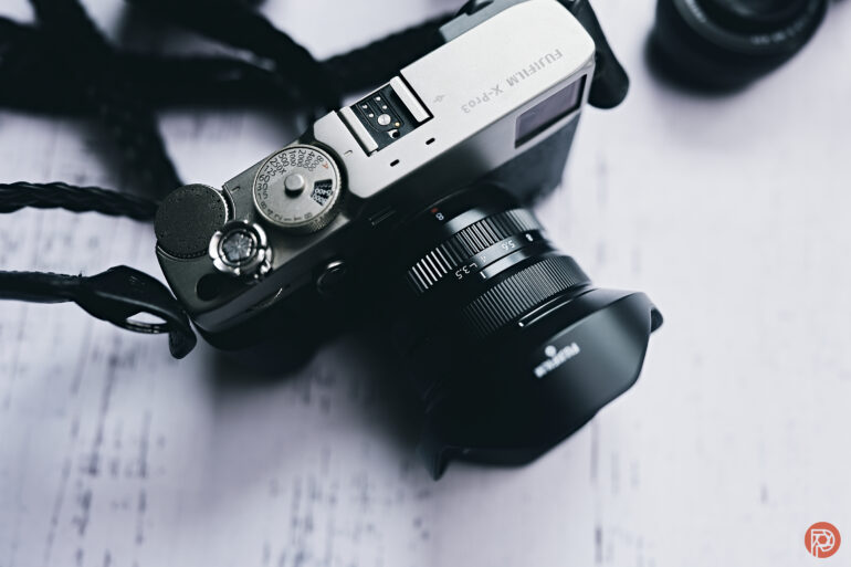 A silver Fujifilm X-Pro3 camera with a lens sits on a white surface, viewed from above.