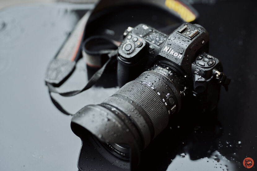 A Nikon camera with a zoom lens is wet with raindrops, resting on a reflective dark surface.