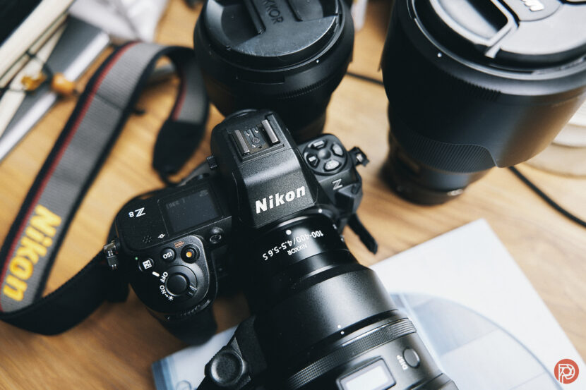 A Nikon camera with attached lens sits on a wooden table beside other camera equipment and accessories.