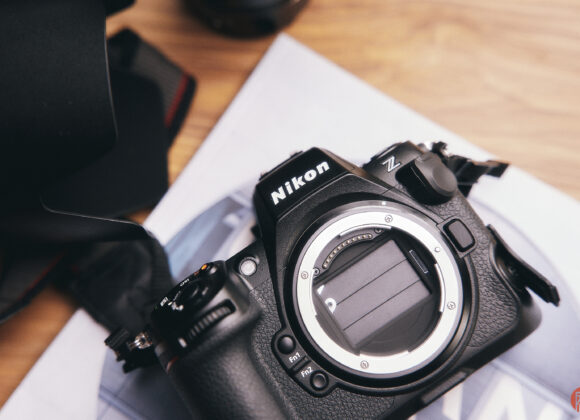 A Nikon Z camera body without a lens sits on a desk, next to a lens hood and some papers.