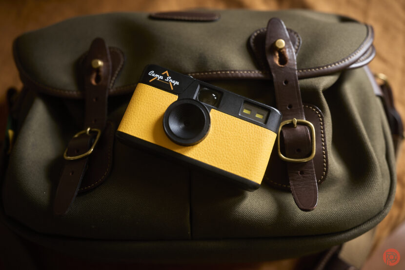 A yellow and black camera placed on an olive green bag with brown straps and buckles.