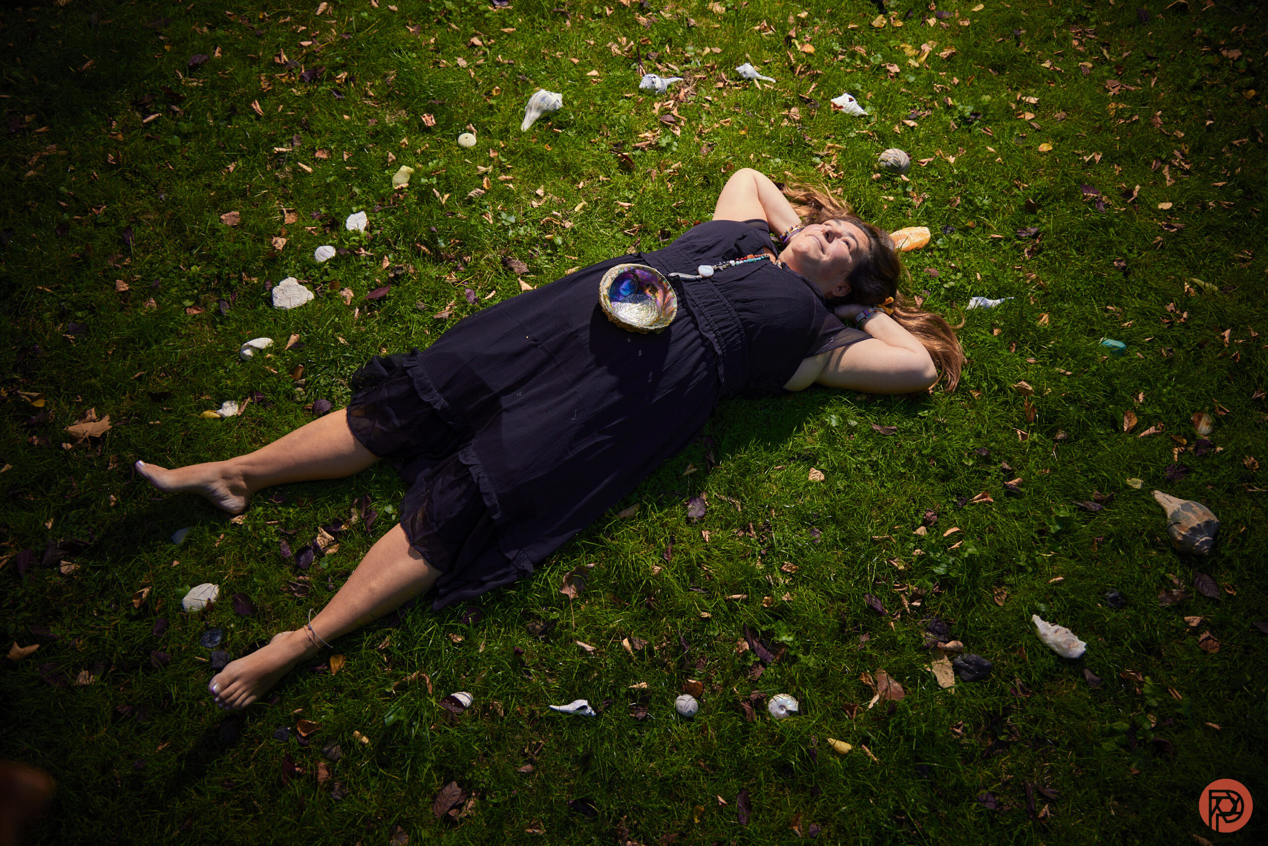 Woman in a black dress lies barefoot on grass, surrounded by scattered stones and leaves.
