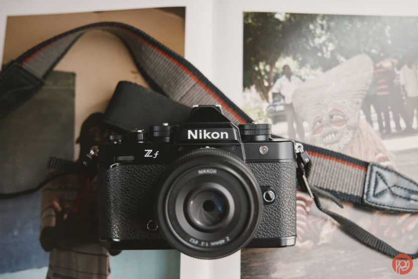 Remember: Win a Nikon Zf With TIPA! A Nikon Z fc camera with a strap placed on an open magazine showing colorful photos.