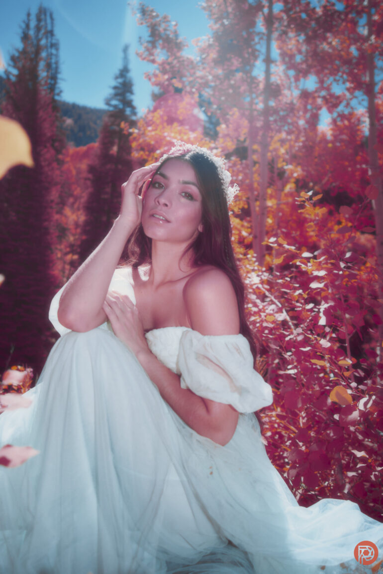 Woman in a white dress sits among vibrant pink autumn foliage under a bright sky, looking at the camera.