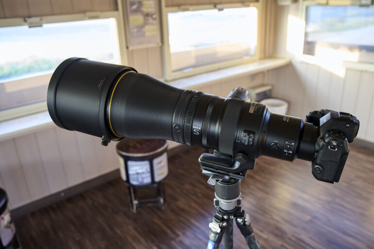 A large telephoto camera lens mounted on a tripod, set up indoors near windows.