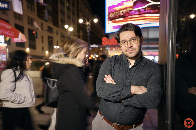 If You're Nothing Without Photoshop, Should You Be Making Photos? Man with glasses leans against reflective surface in a busy city street at night, lights and crowd in background.