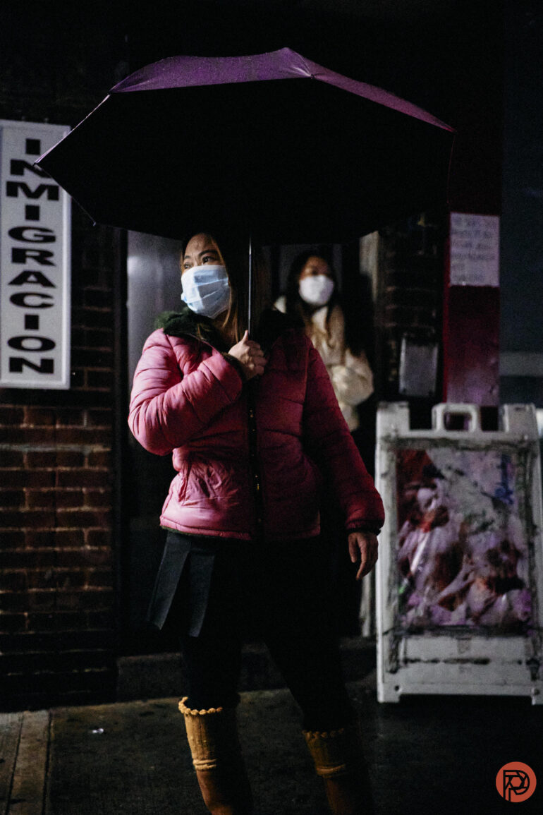 Woman in a pink jacket and mask holds a purple umbrella at night near an Immigracion sign.