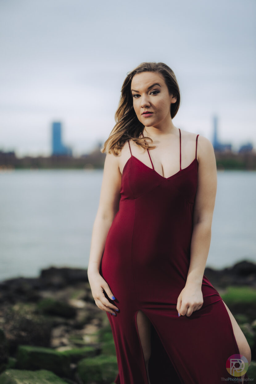 Woman in a burgundy dress poses outdoors by water with a city skyline in the background.