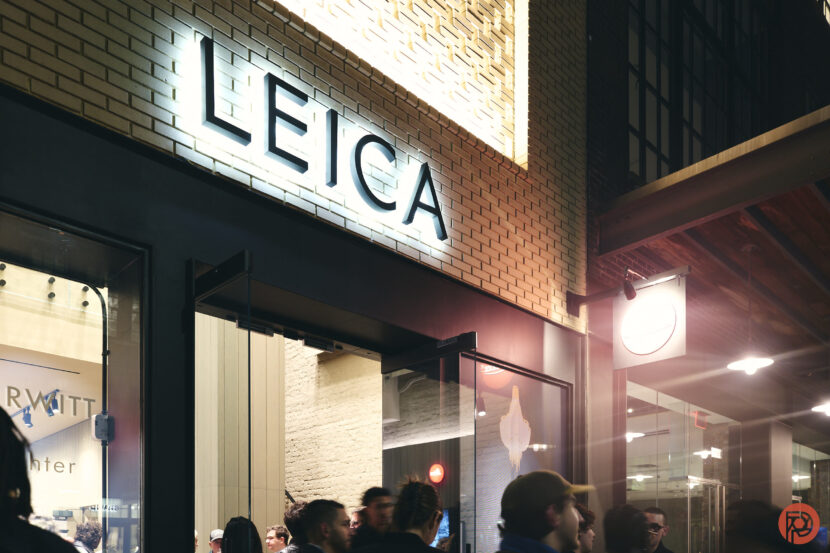 Crowd outside a Leica store at night, with the illuminated Leica sign on a brick wall above the entrance.