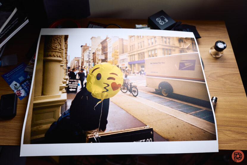 Are Gen Z Printing More Images than Boomers? A photo print on a desk shows a person with a winking emoji balloon face on a city street.
