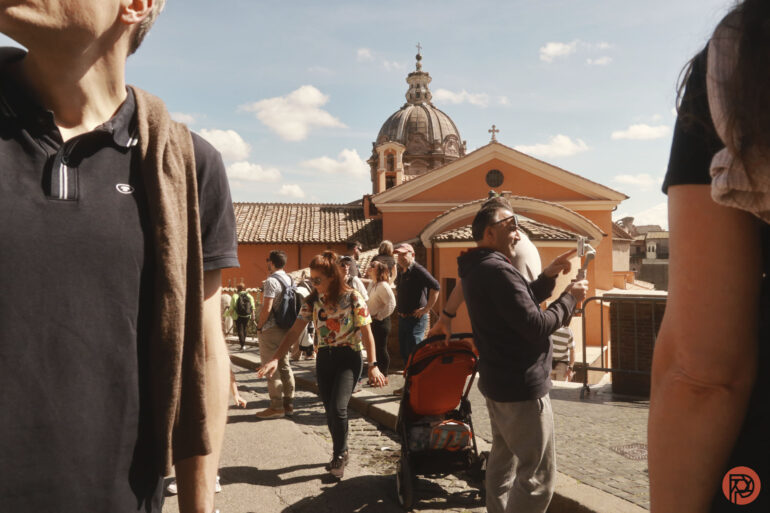 People sightseeing and taking photos in front of historic buildings under a blue sky.