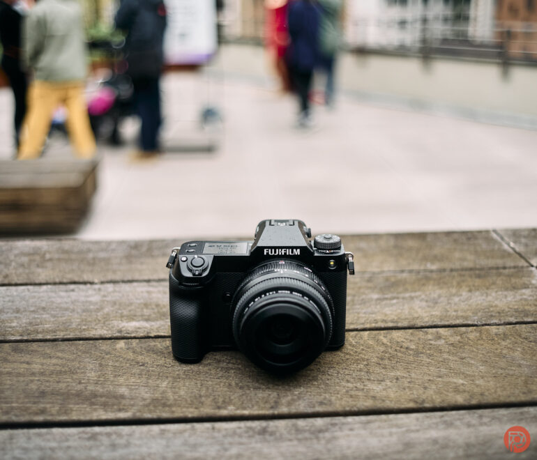 A Fujifilm camera sits on a wooden surface, with a blurred outdoor scene in the background.