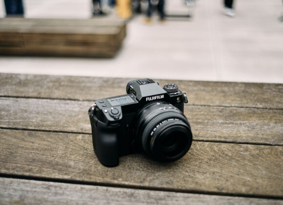 Fujifilm camera on a wooden table with blurred people and outdoor setting in the background.