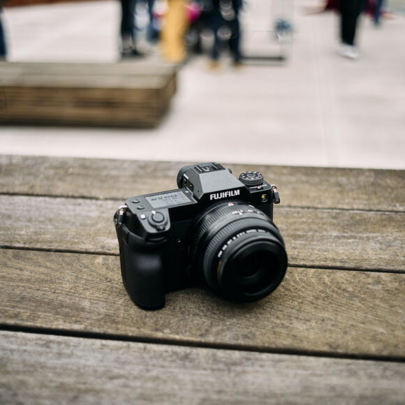 Fujifilm camera on a wooden table with blurred people and outdoor setting in the background.