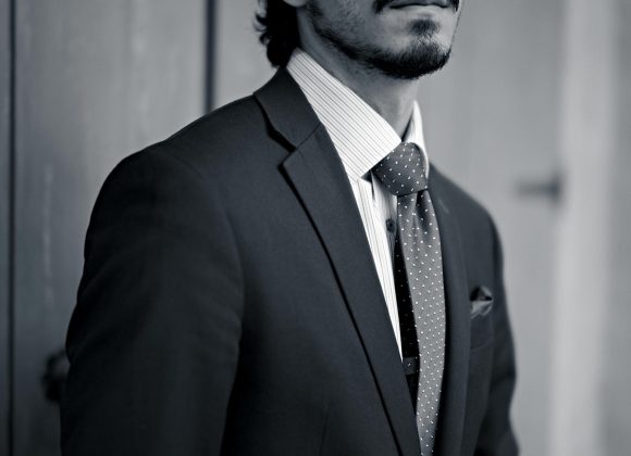 A man in a suit and tie stands in front of wooden panels, looking thoughtfully into the distance.