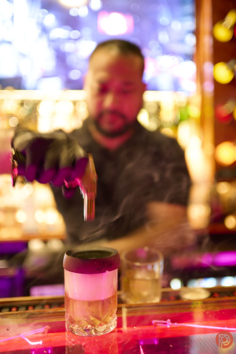 A bartender uses tongs to place a garnish on a smoking cocktail at a vibrant, colorful bar.