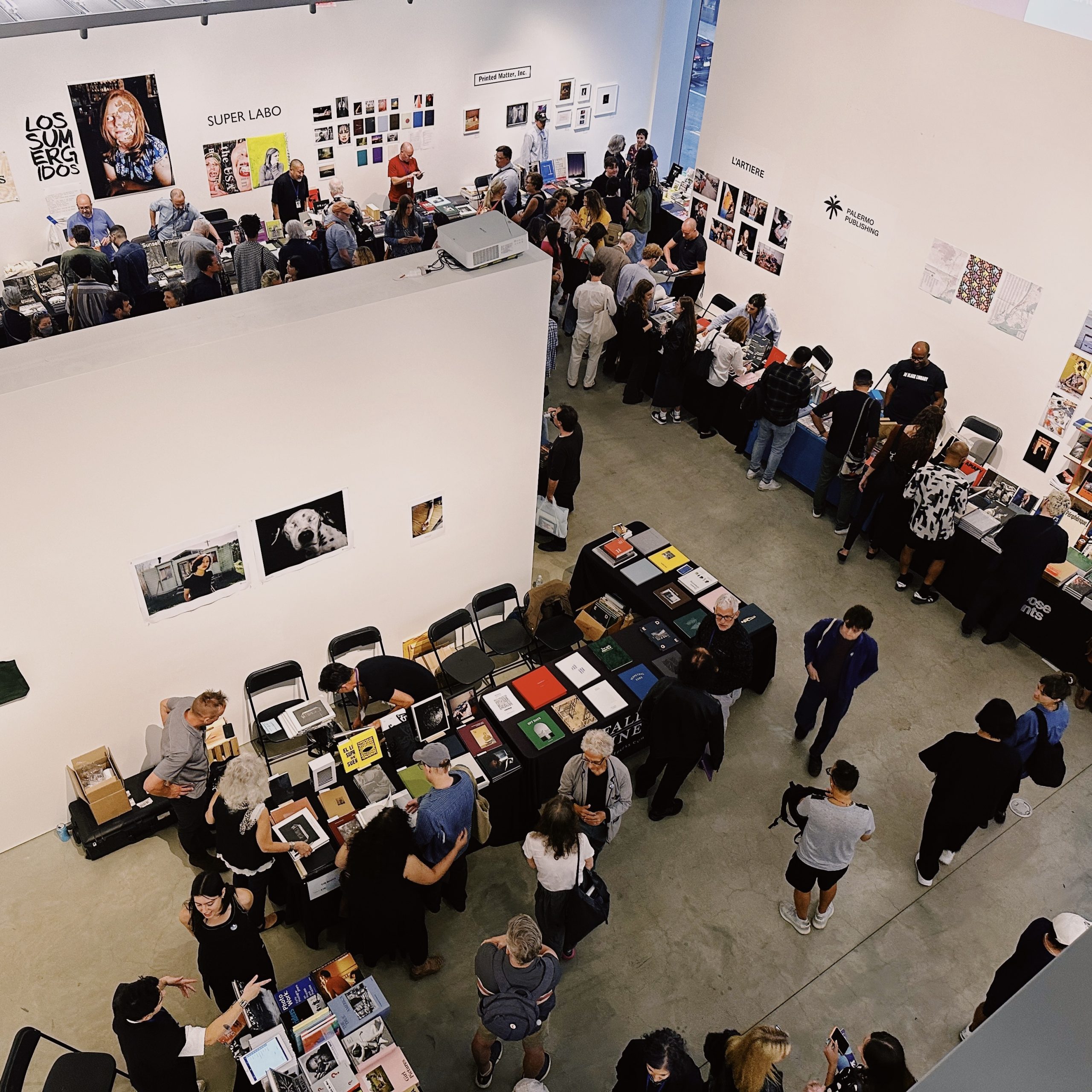 A busy indoor art fair with people browsing tables of books and artworks in a modern gallery space.