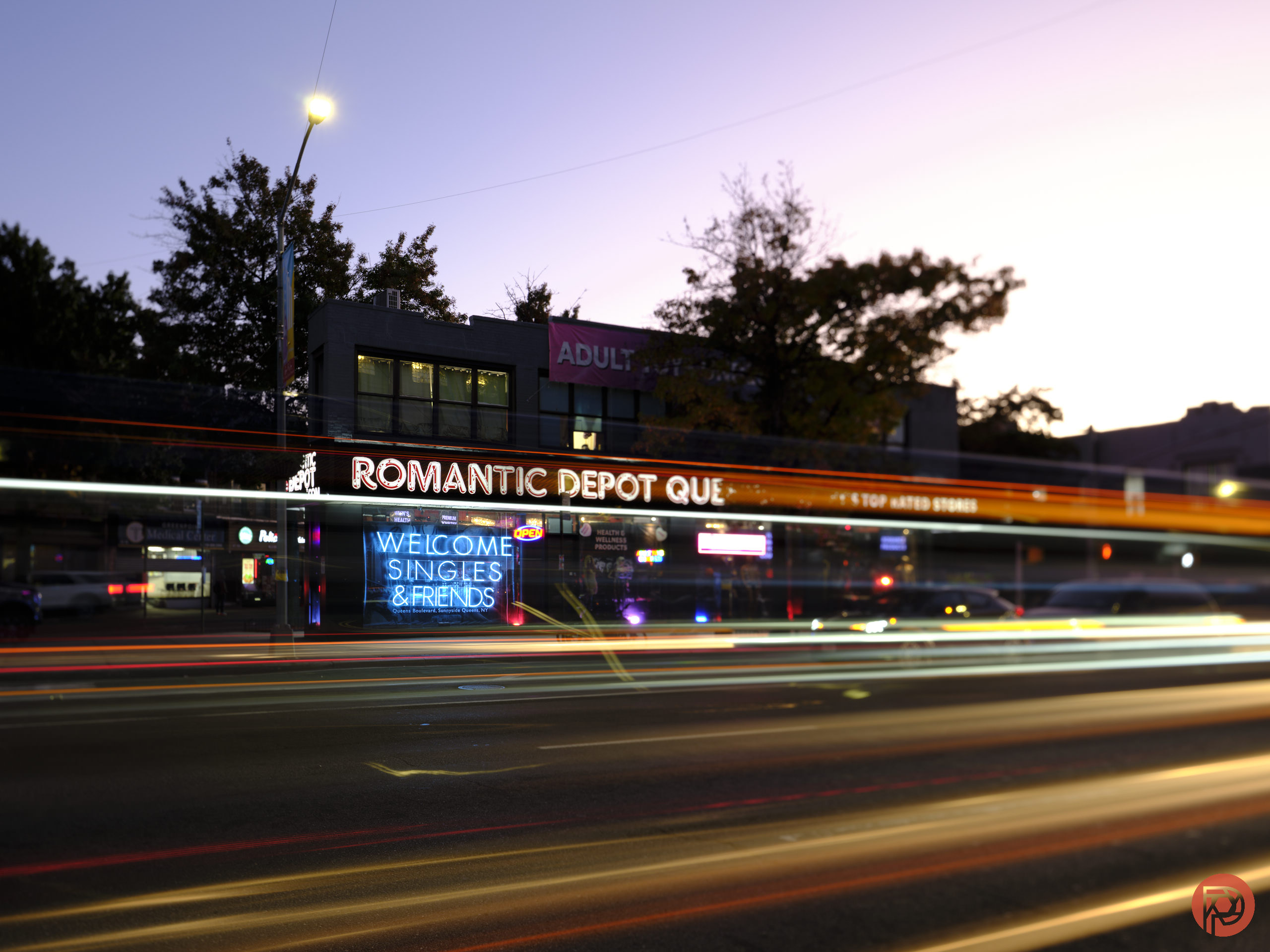 A street view of an adult store with neon signs at dusk and blurred traffic lights passing by.