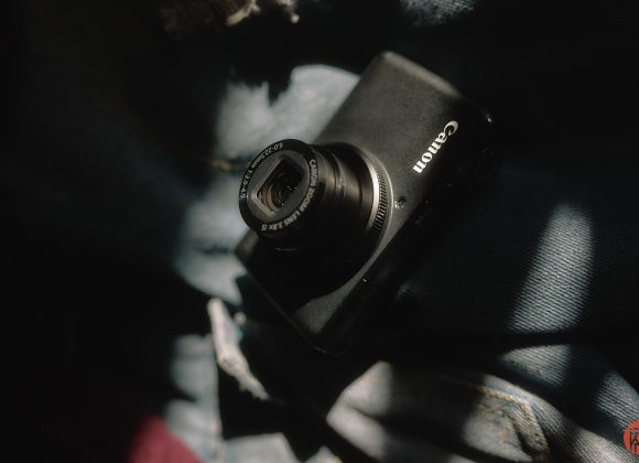 A black Canon digital camera rests on blue denim fabric in soft, dappled sunlight.