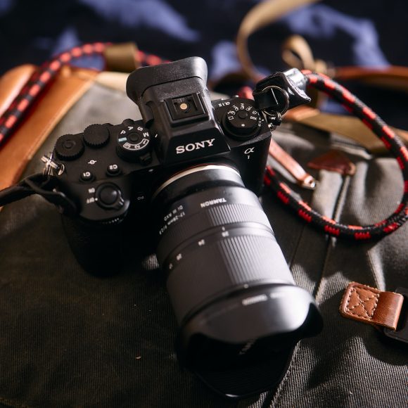 A Sony camera with a large lens rests on a canvas and leather bag in natural light.