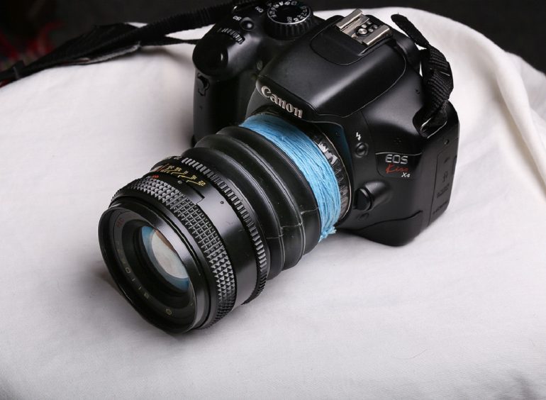 A black Canon EOS camera with a vintage lens attached sits on white fabric, DIY-style, wrapped in blue tape.