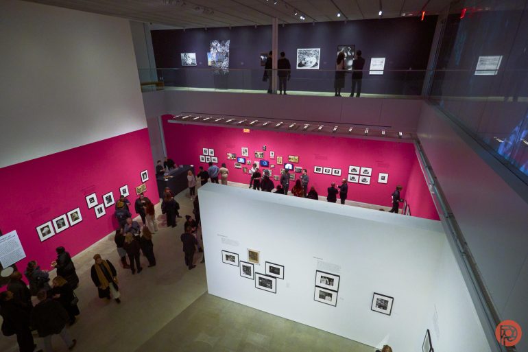 Museum gallery with people viewing framed artworks on vibrant pink and white walls. Two levels are visible.